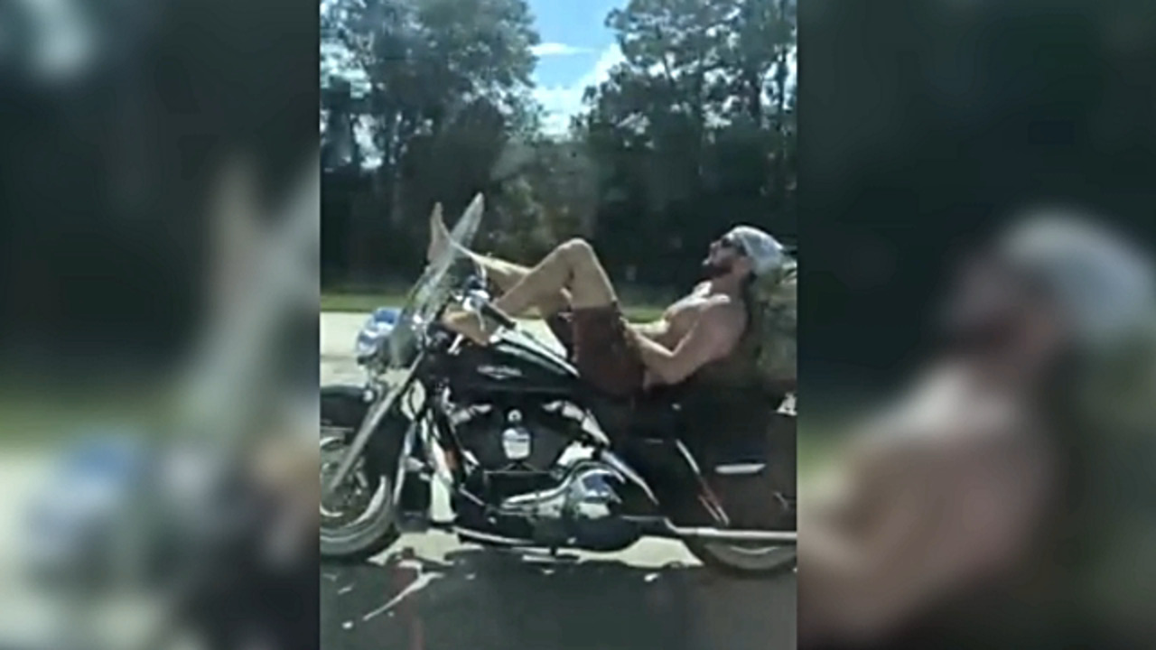 Shirtless man rides motorcycle down highway while lying on his back